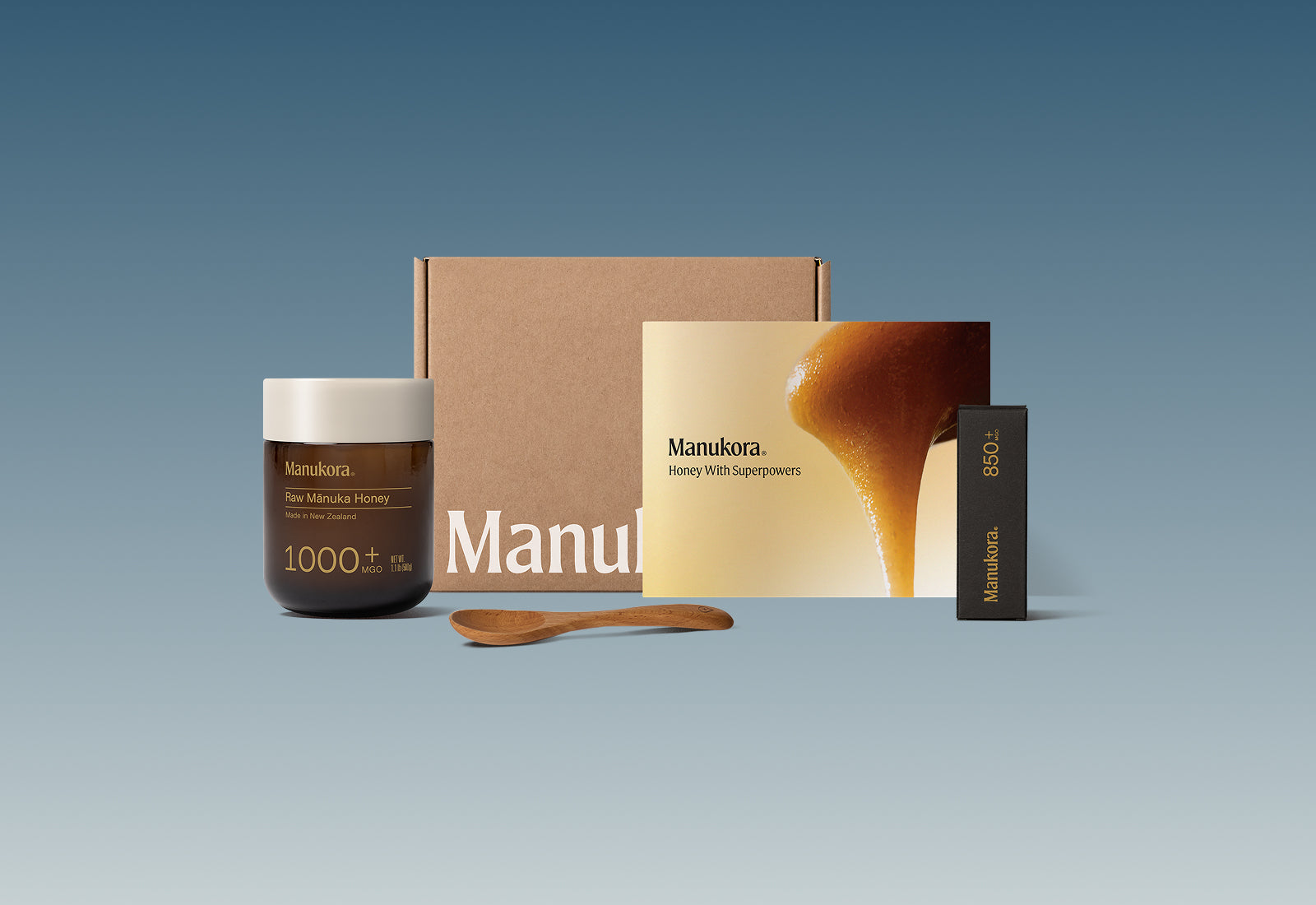 Set of Manukora raw Manuka honey products, including a jar, packaging box, spoon, and promotional card.