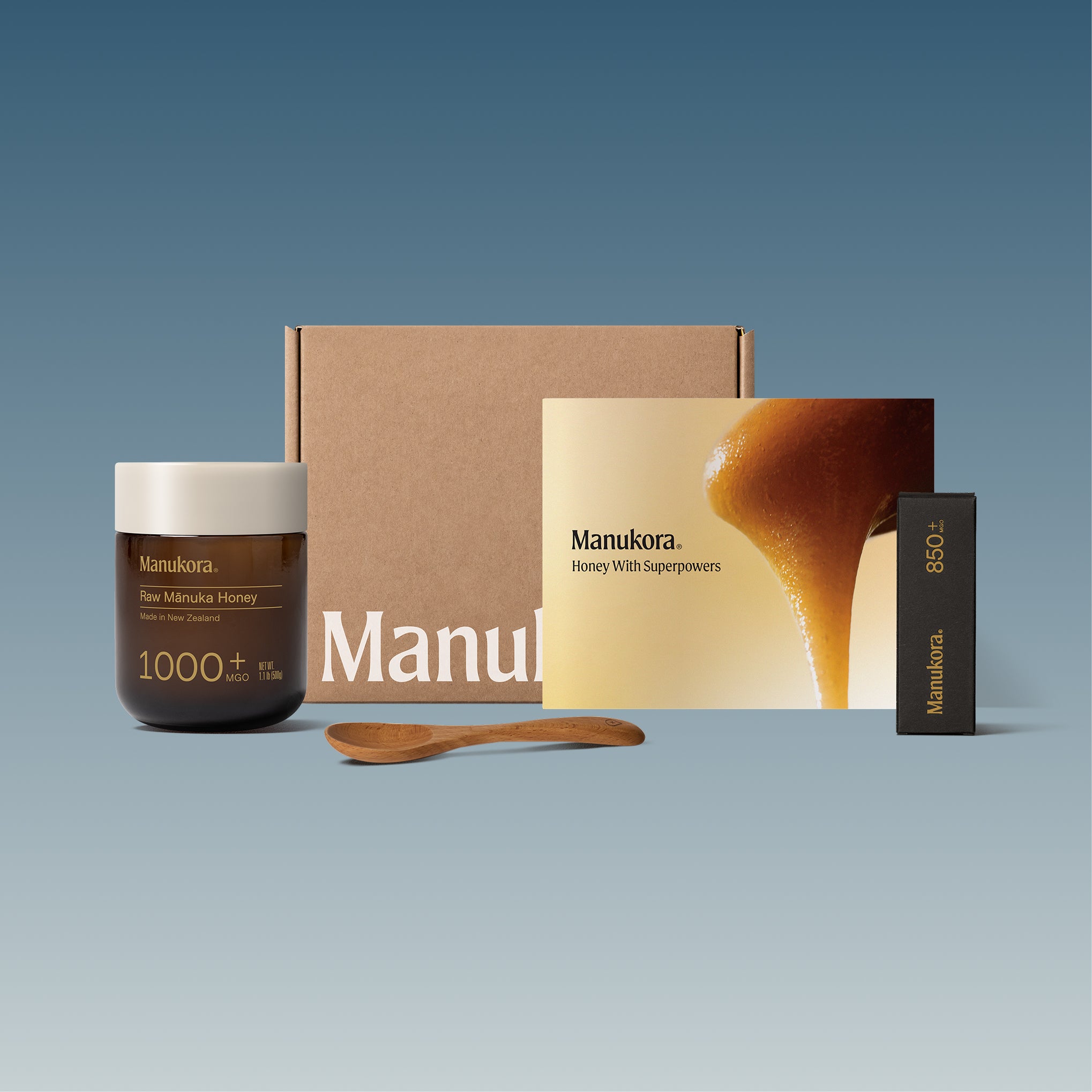 Set of Manukora raw Manuka honey products, including a jar, packaging box, spoon, and promotional card.