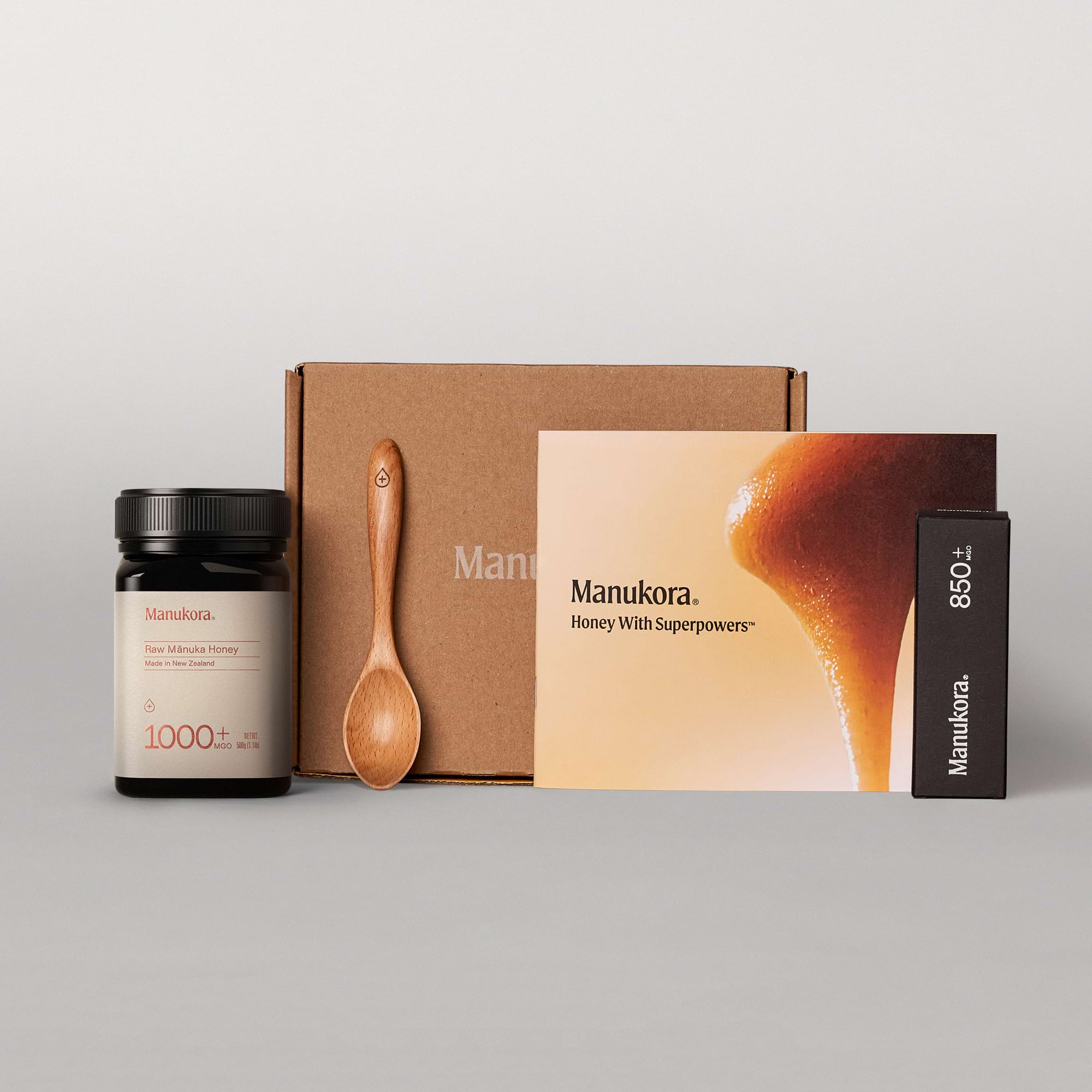 Set of Manukora raw Manuka honey products, including a jar, packaging box, spoon, and promotional card.